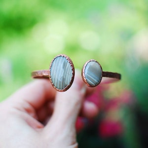 May include: A copper bracelet with two oval-shaped stones set in the metal. The stones are a light gray color with a striated pattern.