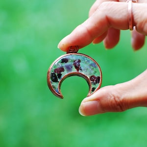 May include: A crescent moon shaped pendant made of copper wire with a clear resin filled with small green, red, and black stones.