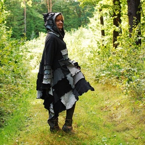 Könnte beinhalten: Ein schwarzer Kapuzenmantel mit mehrschichtigen Patchwork-Details in Grau- und Schwarztönen. Der Umhang hat ein einzigartiges, asymmetrisches Design und wird im Freien in einer Waldumgebung getragen. Die Kapuze des Umhangs ist hochgezogen.