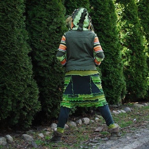 May include: A person wearing a green hooded jacket with striped sleeves, a green and black skirt with layered ruffles, and dark green trousers. The outfit is set against a backdrop of tall, green trees.