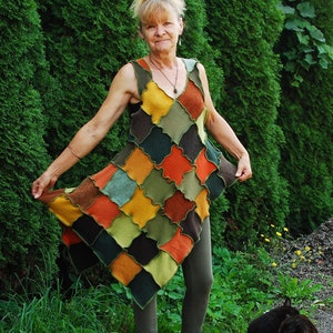 May include: A woman wearing a patchwork dress with a variety of colours, including green, brown, yellow, and orange. She is standing in front of a green hedge and has a black cat sitting in front of her.