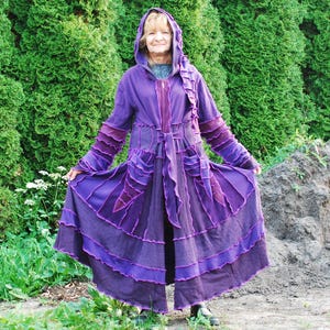 May include: A long, hooded purple coat with layered panels and pockets. The coat features varying shades of purple and a flowing skirt. The person is standing in front of a backdrop of green foliage.