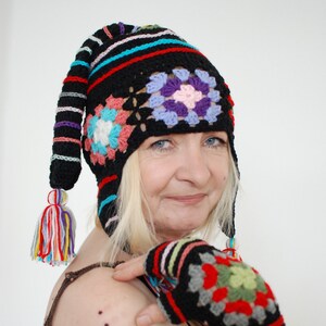 May include: A woman wearing a black crochet hat with colorful stripes and granny squares. The hat has a long, pointed tail with a tassel. She is also wearing a black crochet fingerless glove with colorful stripes and granny squares.
