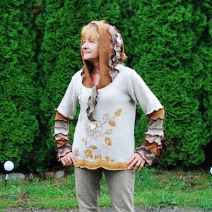 Puede incluir: Una mujer lleva un suéter de punto marrón y blanco con un diseño floral y una capucha marrón. El suéter tiene volantes en las mangas y un ribete marrón alrededor del cuello.