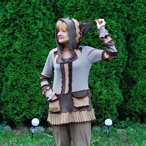 Puede incluir: Una mujer lleva un suéter con capucha marrón y beige con un dobladillo con volantes. El suéter tiene un diseño a rayas marrón y beige y un bolsillo marrón y beige. La mujer está de pie frente a un seto verde.