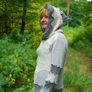 May include: A woman wearing a grey and white hooded sweater with a ruffled trim. The sweater is layered and has a patchwork design. She is standing in a woodland setting.