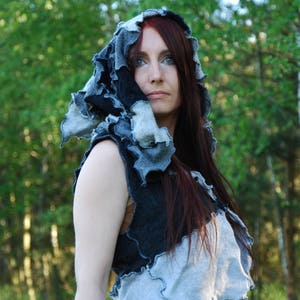 May include: A woman with long, dark red hair wears a hooded vest made of patchwork fabric in shades of grey and black. The vest has a ruffled, asymmetrical hemline and is worn over a brown top.