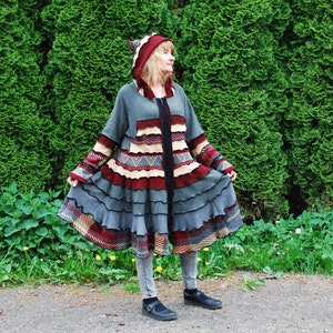 May include: A long, grey coat with a hooded design, featuring a mix of textures and colours. The coat has a ruffled skirt, with accents of burgundy, cream, and patterned details. The person is wearing grey leggings and black shoes.