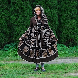 Könnte beinhalten: Ein brauner Kapuzenmantel mit Patchwork-Muster und weißen Verzierungen. Der Mantel hat einen langen, fließenden Rock und wird von einer Person getragen, die vor einer grünen Hecke steht.