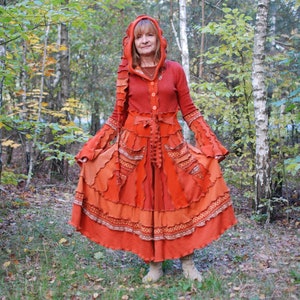 May include: A woman wearing a long, red, hooded dress with a layered, ruffled skirt. The dress is made of a patchwork of different fabrics in shades of orange and brown. The woman is standing in a woodland setting.