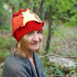 Op de afbeelding: Een kleurrijke, handgemaakte hoed met een uniek ontwerp. De hoed heeft een rode geribbelde band, met een gefranjerde bovenkant in tinten oranje, geel en groen. Een levendig accessoire, perfect om elke outfit op te fleuren.