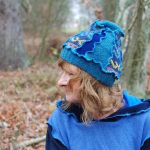 May include: A woman wearing a blue knitted hat with a ruffled edge and a blue hooded sweater. The hat has a yellow and blue pattern.
