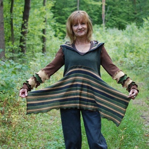 May include: A woman wearing a green, brown and beige striped tunic with long sleeves and a ruffled trim. The tunic has a loose fit and is layered over a green shirt. The woman is standing in a woodland setting.