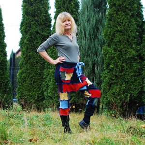 May include: A woman wearing a grey long-sleeved shirt and colourful patchwork trousers stands in front of a row of evergreen trees.