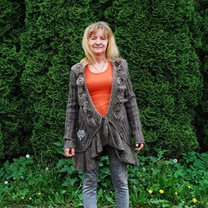 Puede incluir: Una mujer con cabello rubio lleva una chaqueta de punto gris con un suéter estampado marrón y naranja debajo. Está de pie frente a un seto verde.