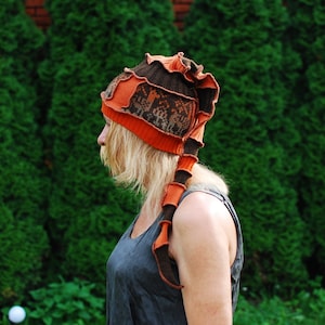 Puede incluir: Una mujer lleva un gorro de punto marrón y naranja con una cola larga. El gorro tiene un diseño único con una mezcla de texturas y colores.