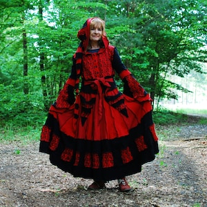 May include: A woman wearing a red and black hooded dress with a ruffled skirt and a black belt. She is standing in a woodland setting.