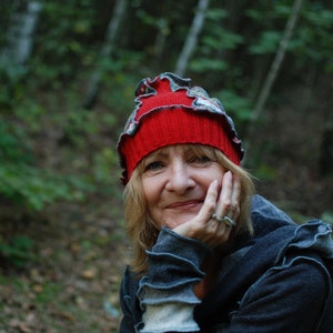 Puede incluir: Una mujer con cabello rubio lleva un gorro de punto rojo y gris con un borde rizado. Está sonriendo y mirando a la cámara. Lleva un suéter gris y tiene un anillo en el dedo.