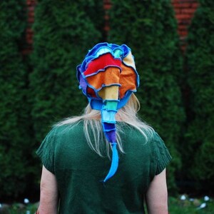 Può includere: Una donna indossa un cappello all'uncinetto colorato con una lunga coda. Il cappello è fatto di diverse tonalità di blu, rosso, arancione, giallo e verde. La donna indossa una camicia verde e ha lunghi capelli biondi.