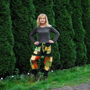 May include: A woman wearing a brown sweater and colorful patchwork pants stands in front of a green hedge.