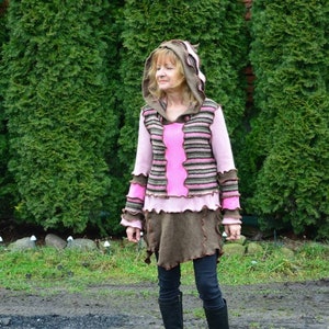 Puede incluir: Una mujer lleva un suéter con capucha a rayas marrones y rosas con un dobladillo marrón con flecos. El suéter tiene un patrón a rayas rosas y marrones y un ribete de volantes rosa. La mujer está de pie frente a una fila de árboles de hoja perenne.