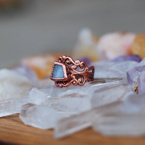 May include: A copper ring featuring a delicate filigree design and a light blue gemstone. The ring is resting on a bed of clear quartz crystals.