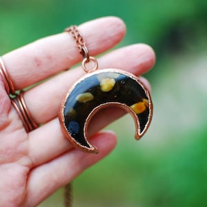 May include: A copper crescent moon pendant with black resin and embedded yellow and white flecks. The pendant is on a copper chain.