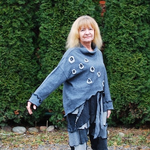 Puede incluir: Una mujer lleva un suéter gris con detalles en blanco y negro. El suéter tiene un diseño único con un dobladillo irregular y un corte holgado. La mujer está de pie frente a un seto verde.