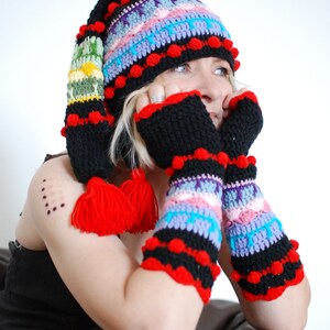 Puede incluir: Una mujer lleva un gorro de ganchillo de colores con pompones y guantes sin dedos a juego. El gorro y los guantes son negros, rojos, azules, verdes y morados.