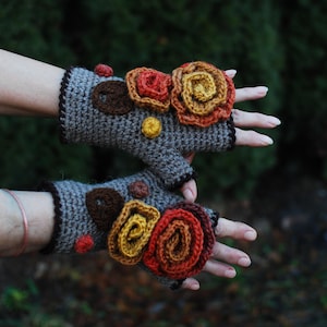Puede incluir: Un par de guantes sin dedos grises con adornos de flores de ganchillo en tonos naranja, amarillo y marrón.