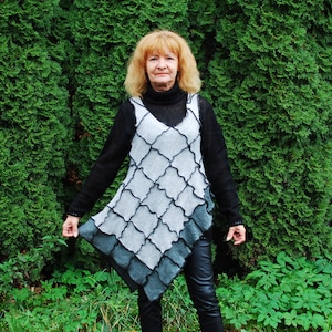 May include: A woman wearing a black long-sleeved shirt with a gray and black patterned tunic over it. She is standing in front of a green hedge.