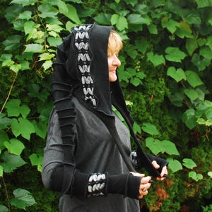 May include: Black hooded scarf with long sleeves and fingerless gloves. The hood has a decorative white and grey pattern. The sleeves and gloves have a layered design. The person is wearing a grey long-sleeved shirt.
