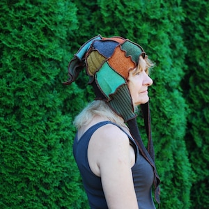 May include: A woman wearing a colorful, patchwork hat with a long, brown tassel. The hat is made of different fabrics and colors, including green, blue, orange, and brown. The woman is standing in front of a green hedge.