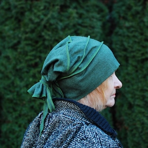 Puede incluir: Un gorro verde tejido con un diseño único, con detalles verticales y un cierre anudado. El gorro es de un tono verde intenso. La persona lleva una chaqueta con estampado en blanco y negro.