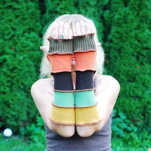 Könnte beinhalten: Eine Person trägt ein Paar bunte gestreifte Fingerlose Handschuhe. Die Handschuhe sind aus weichem, gestricktem Stoff und haben ein einzigartiges, mehrschichtiges Design. Die Handschuhe sind eine Mischung aus Grün, Orange, Schwarz und Braun.