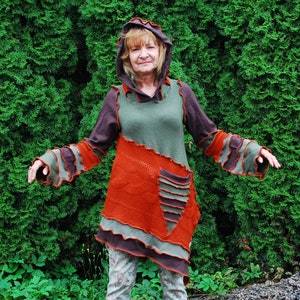 Puede incluir: Una mujer lleva una túnica con capucha multicolor con un diseño de patchwork. La túnica está hecha de una tela suave y de punto y presenta una variedad de colores, incluyendo marrón, verde y naranja. La mujer está de pie frente a un seto verde.