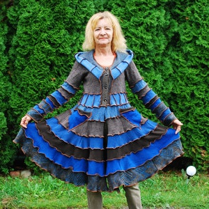 May include: A woman wearing a blue and brown layered jacket with a ruffled hem. The jacket is made of different fabrics and has a unique, bohemian style.