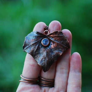 Puede incluir: Un colgante de hoja de cobre con una piedra preciosa púrpura en el centro. El colgante se sostiene en una mano con varios anillos de cobre en los dedos.