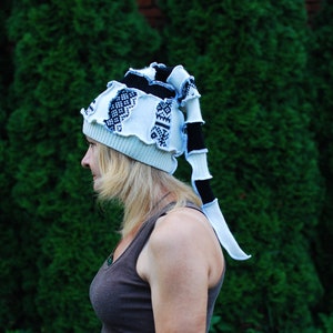 May include: A quirky, handmade hat with a long, striped tail. The hat has a pale blue base with white and black details, including patterned patches. The design is unique and eye-catching, ideal for adding a touch of fun to any outfit.