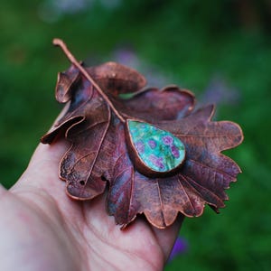 Op de afbeelding: Een handgemaakt koperen blad met een druppelvormige hanger. De hanger heeft een turquoise en robijnrode steen, gezet in een koperen bezel. Het blad is diep roodbruin van kleur, met ingewikkelde adering.