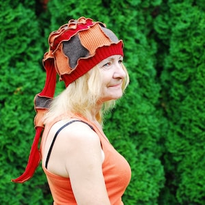 Puede incluir: Una mujer con un sombrero de colores con un largo rabo. El sombrero está hecho de tela roja, naranja, marrón y negra. La mujer lleva una camiseta de tirantes naranja brillante.