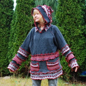 Puede incluir: Un suéter gris con capucha con ribetes rojos y blancos. El suéter tiene un diseño con volantes y bolsillos. El suéter lo lleva puesto una persona de pie frente a una fila de árboles de hoja perenne.