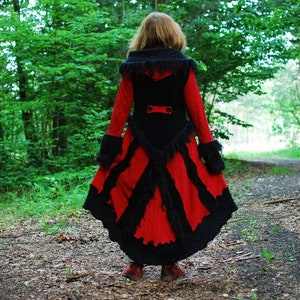 May include: A woman wearing a long, red and black coat with a fur trim. The coat has a fitted bodice and a flowing skirt. The woman is standing in a woodland setting.