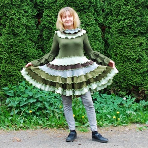 Pode incluir: Uma mulher vestindo um vestido verde, marrom e branco com camadas de babados, mangas compridas e gola alta. Ela está em pé na frente de uma sebe verde.