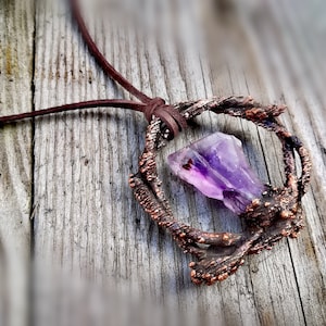 Puede incluir: Un colgante de cristal de amatista púrpura engastado en un marco de alambre de cobre, que cuelga de un cordón de cuero marrón.