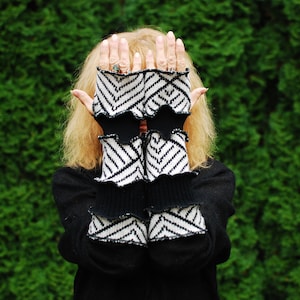 Puede incluir: Guantes sin dedos de punto blanco y negro con un patrón de chevrón. Los guantes tienen un borde con volantes y se usan sobre una camiseta negra de manga larga. El fondo es un seto verde.