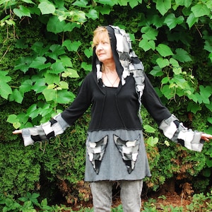 Puede incluir: Una parte superior con capucha negra con detalles de patchwork en la capucha, las mangas y la falda. Las mangas y la capucha presentan un patrón de cuadros en blanco, negro y gris. La falda tiene dos bolsillos de patchwork grises y negros. La persona está de pie frente a un fondo verde frondoso.