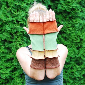 Könnte beinhalten: Ein Paar bunte Fingerlose Handschuhe mit einem gestreiften Muster in Braun-, Grün- und Beigetönen. Die Handschuhe sind aus weichem, gestricktem Stoff.