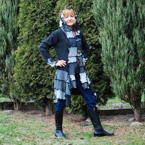 May include: A woman wearing a long, hooded, patchwork coat with a blue and gray color scheme. The coat has a frayed, distressed look and is layered with different fabrics. She is wearing blue jeans and black boots.