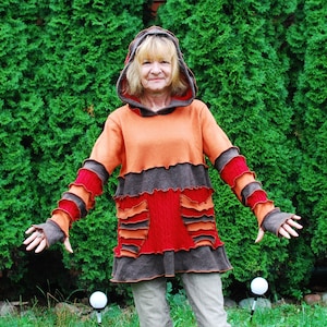 Puede incluir: Una mujer lleva un suéter con capucha multicolor con un diseño en capas y con volantes. El suéter es marrón, naranja y rojo. La mujer está de pie frente a un seto verde.